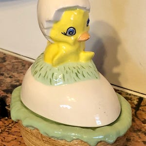 May include: A ceramic Easter-themed figurine of a yellow chick hatching from a white egg. The chick has blue eyes and an orange beak. The egg sits on a light green base, and the entire piece is made of ceramic.