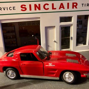 May include: A red model car with silver rims and a black antenna is parked in front of a Sinclair Tires service station. The service station has white walls and green trim, with the Sinclair Tires logo displayed.