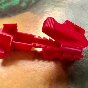 Può includere: Un componente di giocattolo in plastica rosso brillante con un design geometrico complesso. Il pezzo presenta più sezioni rettangolari e angolate, con una parte incernierata e una serie di sporgenze a forma di dente lungo un bordo. Il giocattolo sembra far parte di un set di costruzione più grande.