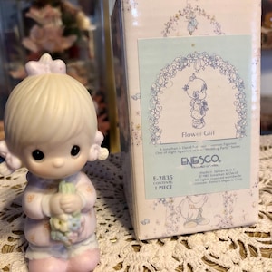 May include: A porcelain figurine of a girl holding flowers, part of the "Wedding Party Series." The figurine is in a pink dress and has a bow in her hair. The box reads "Flower Girl" and "ENESCO."