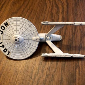 Könnte beinhalten: Ein weißes Modell des Raumschiffs Enterprise mit dem Text "NCC-1701" auf der Untertassensektion. Das Modell hat ein detailliertes Design mit Gondeln und einem Sekundärrumpf. Das Spielzeug ist aus Kunststoff und liegt auf einer Holzoberfläche.