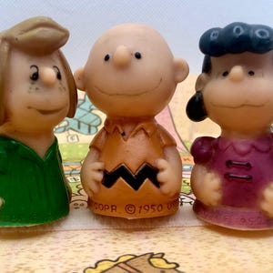 May include: Three vintage Peanuts character figurines: Sally, Charlie Brown, and Lucy. Sally wears a green dress, Charlie Brown has an orange shirt, and Lucy is in a purple dress. The figurines are on a patterned surface.