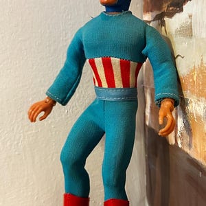 May include: A vintage Captain America action figure. The figure is wearing a blue jumpsuit with a red and white striped design on the chest, a blue helmet with the letter "A", and red boots. The figure is approximately 12 inches tall.