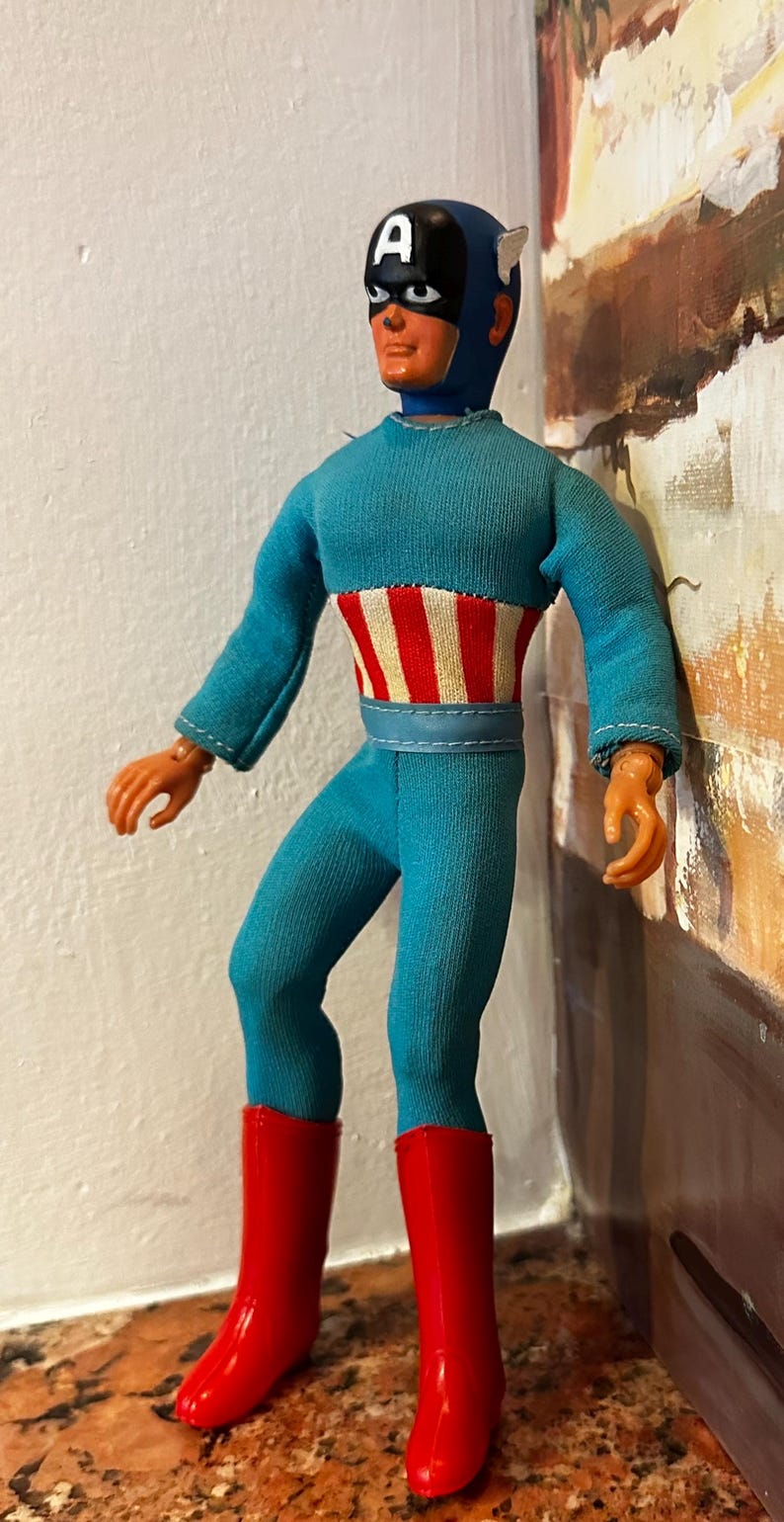 May include: A vintage Captain America action figure. The figure is wearing a blue jumpsuit with a red and white striped design on the chest, a blue helmet with the letter "A", and red boots. The figure is approximately 12 inches tall.