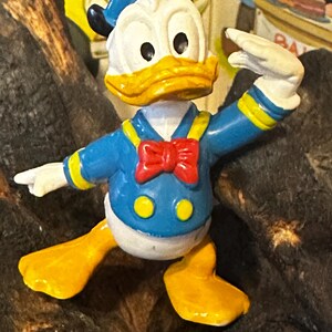 May include: A colorful Donald Duck figurine. The figure is wearing a blue sailor shirt with yellow accents, a red bow tie, and a blue sailor hat. The duck has a yellow beak and feet, and white gloves. The figurine is posed with one arm raised.
