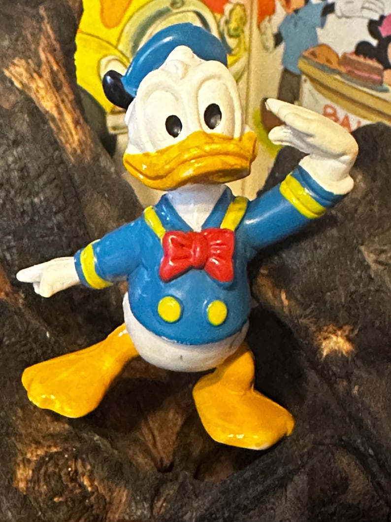 May include: A colorful Donald Duck figurine. The figure is wearing a blue sailor shirt with yellow accents, a red bow tie, and a blue sailor hat. The duck has a yellow beak and feet, and white gloves. The figurine is posed with one arm raised.
