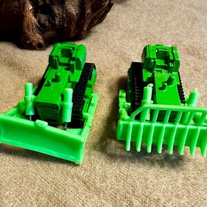 May include: Two bright green toy bulldozers with black treads are positioned side-by-side. The bulldozers have a blade in the front and a protective grill. The toys are on a beige surface.