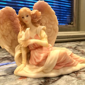 May include: A seated angel figurine with large, detailed wings. The angel wears a flowing pink and cream-colored gown and holds a dove. The figurine is made of a light-colored material with subtle shading.