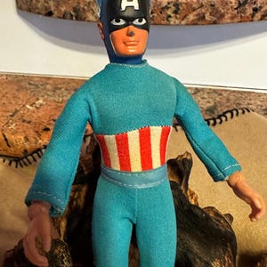 May include: A vintage Captain America action figure. The figure is wearing a blue jumpsuit, red boots, and a black mask with a white "A" on the forehead. The chest features a red and white striped design.