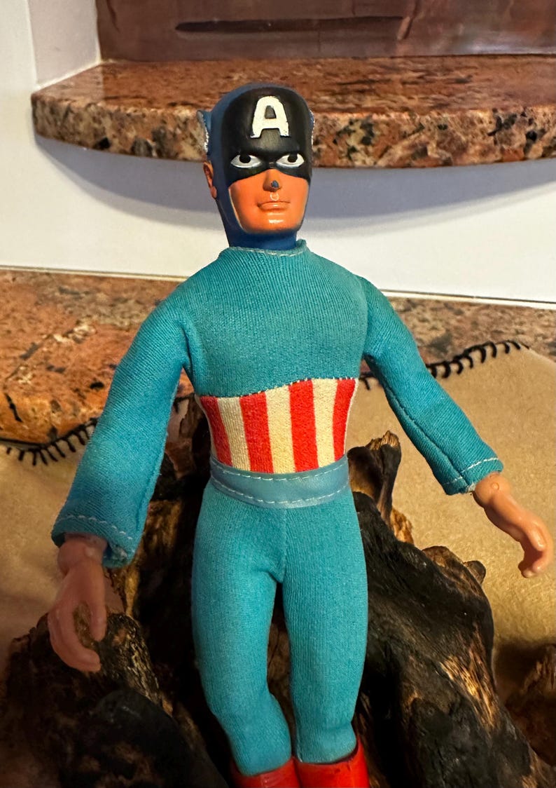May include: A vintage Captain America action figure. The figure is wearing a blue jumpsuit, red boots, and a black mask with a white "A" on the forehead. The chest features a red and white striped design.