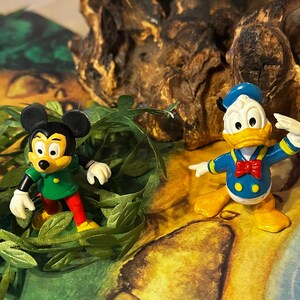 May include: Two Disney character figurines: Mickey Mouse in a green shirt and red pants, and Donald Duck in a blue sailor suit with a red bow tie. The figures are set against a backdrop of greenery and a textured, brown surface.