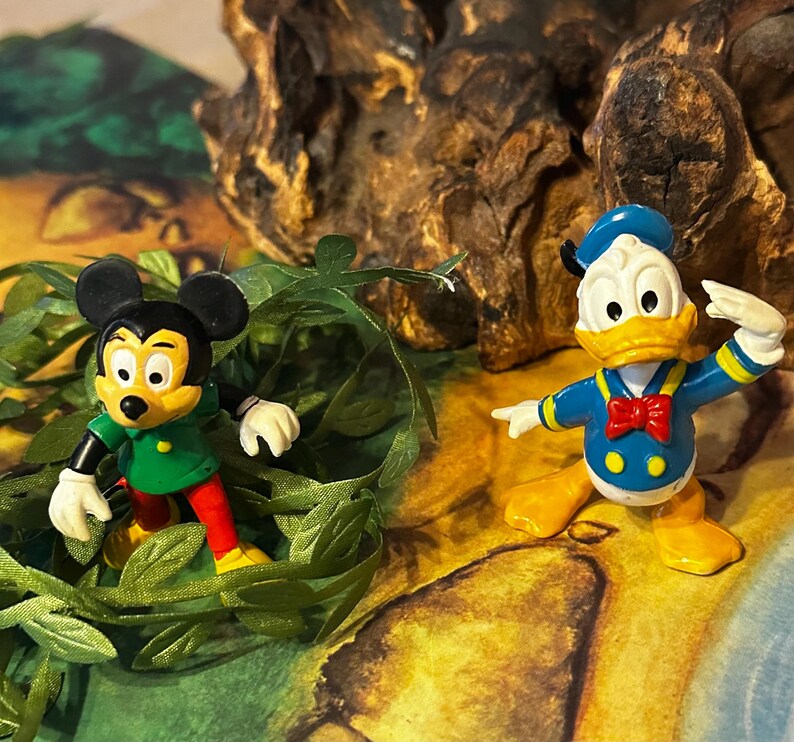 May include: Two Disney character figurines: Mickey Mouse in a green shirt and red pants, and Donald Duck in a blue sailor suit with a red bow tie. The figures are set against a backdrop of greenery and a textured, brown surface.