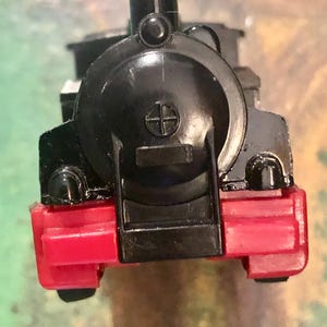 May include: A black toy train with red detailing. The front of the train has a red bumper and a black grill. The train has a round, black smokestack and a circular emblem on the front.