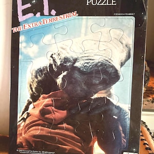 May include: A 15-piece E.T. the Extra-Terrestrial puzzle. The puzzle features a close-up of E.T. and a person in a red jacket. The puzzle box is black with the movie title in light blue and purple.