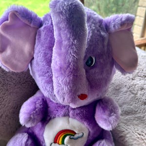 May include: A plush purple elephant toy with large ears and a trunk. The elephant has a rainbow and cloud patch on its chest. The toy has blue eyes and a small red nose. The elephant is sitting on a soft, gray surface.