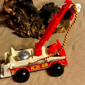 May include: A vintage Fisher-Price toy fire engine, featuring a red crane and accents. The toy has black wheels, a silver bell, and a red and yellow rope. The side of the truck reads "Fisher Price Fire Engine 1".