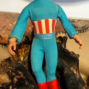 May include: A vintage Captain America action figure. The figure is wearing a blue jumpsuit with a red, white, and blue striped design on the chest. The figure has red boots and a blue mask with a white "A" on the forehead.