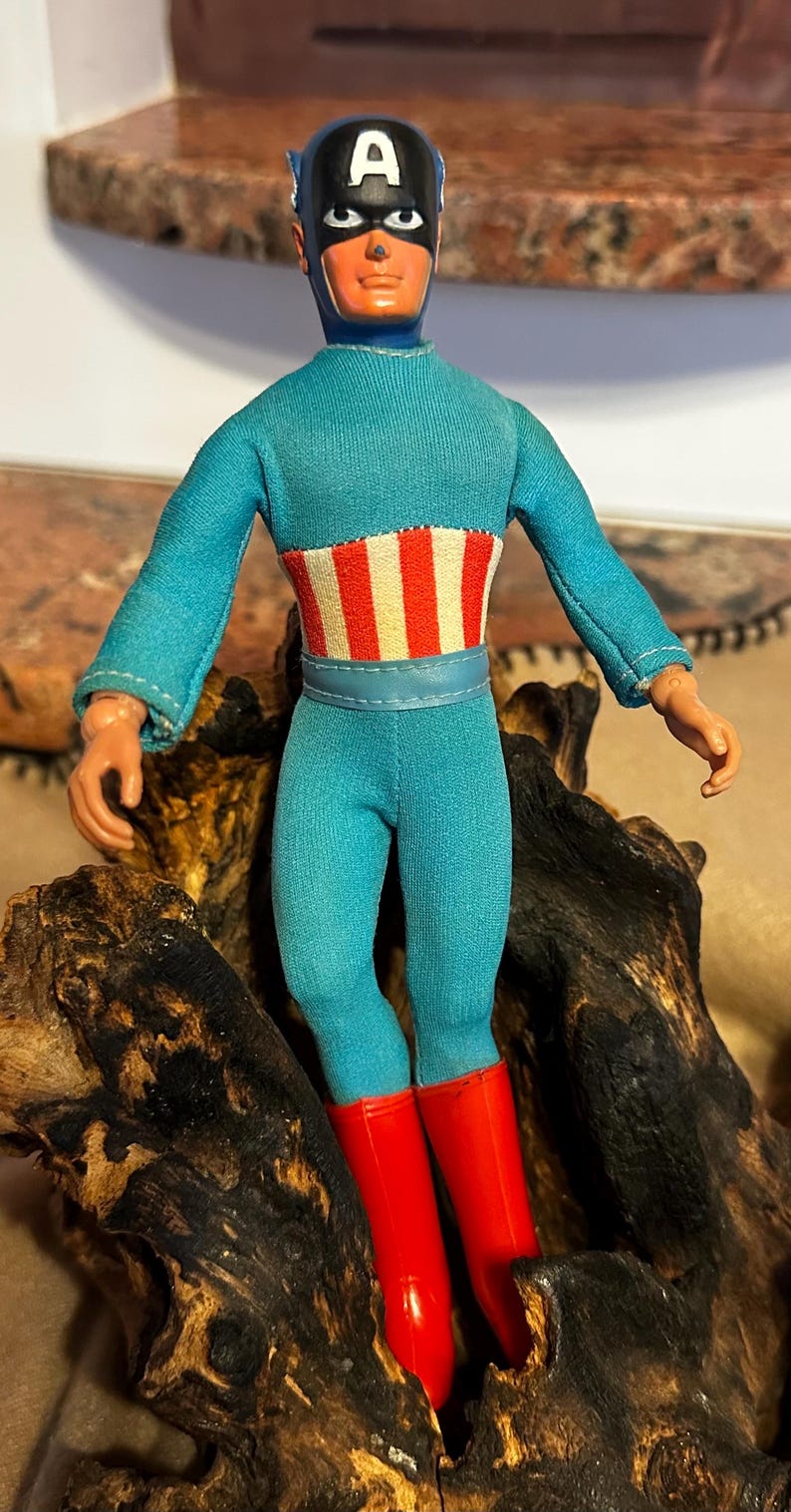 May include: A vintage Captain America action figure. The figure is wearing a blue jumpsuit with a red, white, and blue striped design on the chest. The figure has red boots and a blue mask with a white "A" on the forehead.