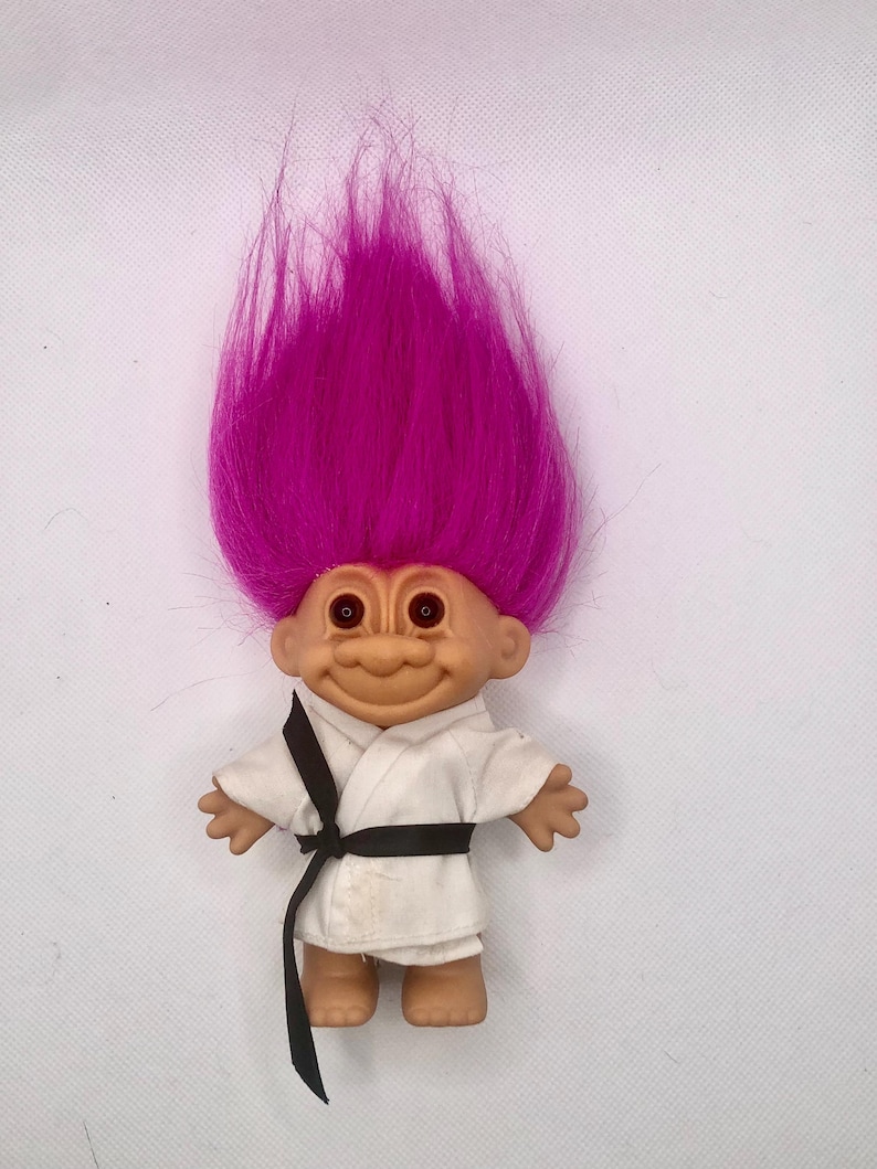 May include: A vintage troll doll with a smiling face and bright pink hair. The doll is wearing a white karate uniform with a black belt. The troll has brown eyes and is standing upright.
