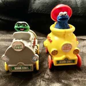Könnte beinhalten: Zwei Spielzeugfahrzeuge mit Charakteren aus der Sesamstraße. Eines ist grau mit Oscar dem Miesepeter und dem Text "I LOVE TRASH". Das andere ist gelb mit Krümelmonster und dem Text "I LOVE COOKIES". Beide haben "Sesame Street" vorne.