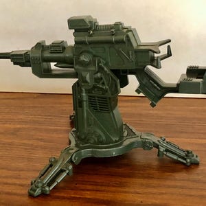 May include: A dark green toy gun with a tripod base. The toy gun has a long barrel and a detailed design, including a sight and a seat-like structure. The base has three legs. The toy is made of plastic.