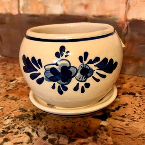 May include: Cream-colored ceramic planter with a matching saucer. The planter features a blue floral design and a blue band around the rim. The round planter is suitable for small plants and adds a touch of rustic charm.