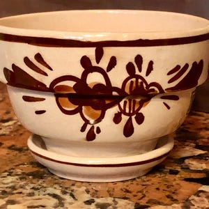 May include: Cream-colored ceramic planter with a matching saucer. The planter features a dark brown floral design and a dark brown band around the rim. The design is repeated on the saucer.