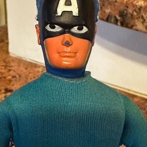May include: A vintage Captain America action figure. The figure has a blue and red striped torso, a blue sweater, and a black mask with a white "A" on the forehead. The face is orange with black eyes.