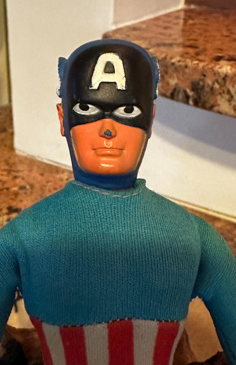 May include: A vintage Captain America action figure. The figure has a blue and red striped torso, a blue sweater, and a black mask with a white "A" on the forehead. The face is orange with black eyes.