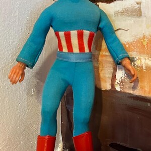 May include: A vintage Captain America action figure. The figure is wearing a blue jumpsuit with a white and red striped midsection and red boots. The figure has a black mask with the letter "A" on the forehead.