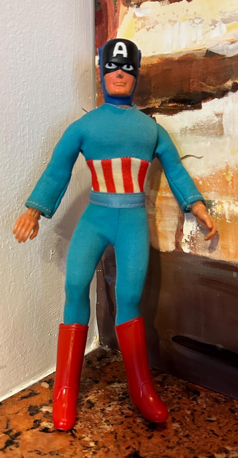 May include: A vintage Captain America action figure. The figure is wearing a blue jumpsuit with a white and red striped midsection and red boots. The figure has a black mask with the letter "A" on the forehead.