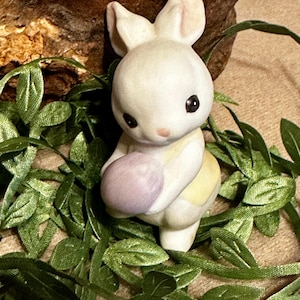 May include: A ceramic Easter bunny figurine, holding a purple egg. The bunny is white with black eyes and a pink nose. It is set against a backdrop of green leaves and a brown wooden element, creating a spring-themed display.