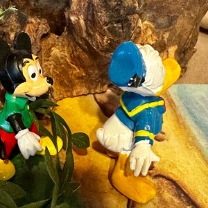 May include: A figurine featuring Mickey Mouse and Donald Duck. Mickey Mouse is wearing a green shirt and red shorts. Donald Duck is wearing a blue sailor suit and hat. The background includes a textured, brown surface and green foliage.
