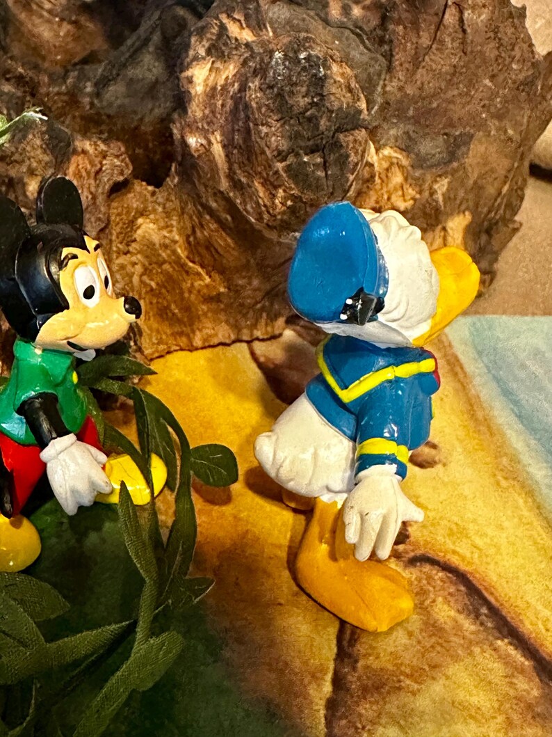 May include: A figurine featuring Mickey Mouse and Donald Duck. Mickey Mouse is wearing a green shirt and red shorts. Donald Duck is wearing a blue sailor suit and hat. The background includes a textured, brown surface and green foliage.