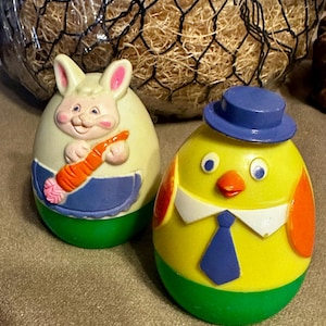 May include: Two Easter-themed egg-shaped figurines. One is a bunny holding a carrot, wearing a blue apron. The other is a yellow chick wearing a blue hat and tie. Both have green bases.