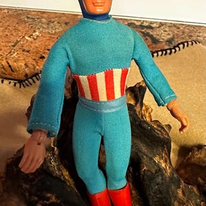 May include: A vintage Captain America action figure. The doll is wearing a blue jumpsuit with a red and white striped midsection, red boots, and a black mask with a white "A" on the forehead. The figure is posed on a piece of wood.
