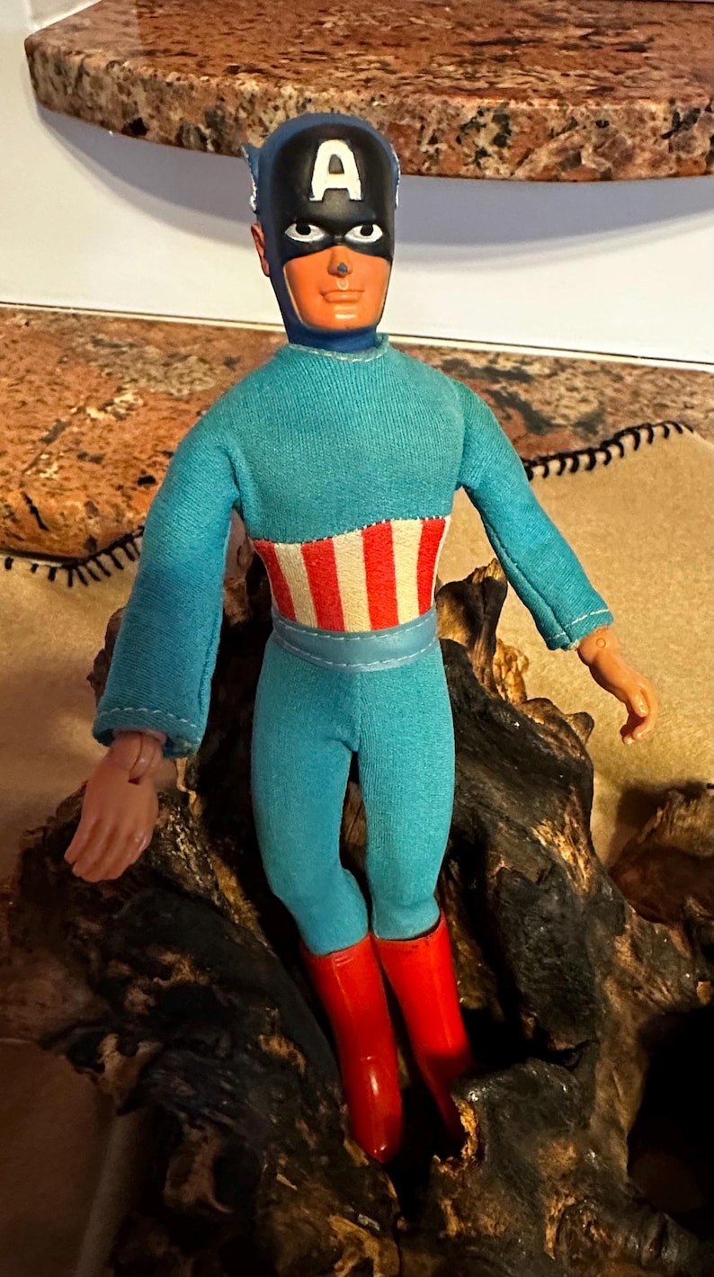 May include: A vintage Captain America action figure. The doll is wearing a blue jumpsuit with a red and white striped midsection, red boots, and a black mask with a white "A" on the forehead. The figure is posed on a piece of wood.