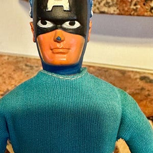 May include: A vintage Captain America action figure. The figure has a blue and white striped torso, a turquoise sweater, and a black mask with a white "A" on the forehead. The figure's face is orange with white eyes and a small nose.