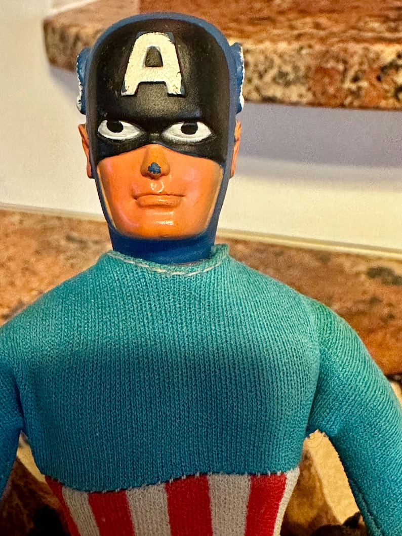 May include: A vintage Captain America action figure. The figure has a blue and white striped torso, a turquoise sweater, and a black mask with a white "A" on the forehead. The figure's face is orange with white eyes and a small nose.