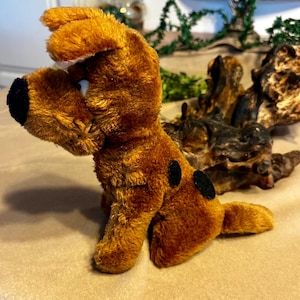 May include: A plush brown dog toy with a black nose and two black spots on its side. The toy is sitting on a beige surface. The dog is a cartoon character.