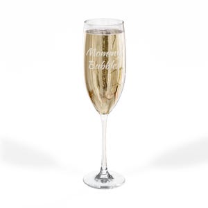 May include: A champagne flute filled with a bubbly, golden liquid. The glass has a slender stem and a wide bowl, with the words "Mommy Bubbles" etched on the front. The glass is set against a white background.