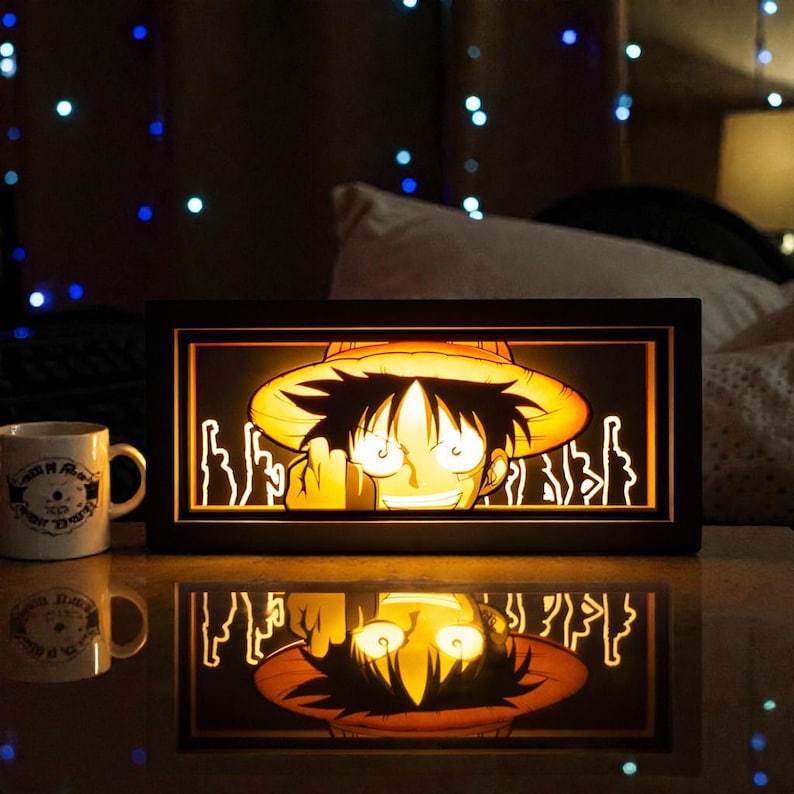 Pu&ograve; includere: Una scatola luminosa con cornice nera raffigurante un personaggio dei cartoni animati con un cappello di paglia. Il volto del personaggio &egrave; illuminato, con un bagliore giallo. A sinistra c'&egrave; una tazza bianca con un disegno.