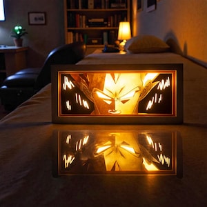 Pu&ograve; includere: Un'opera d'arte rettangolare e illuminata con un personaggio della serie anime Dragon Ball Z. L'opera &egrave; racchiusa in una cornice scura, con il volto e i capelli del personaggio illuminati da una calda luce dorata. L'opera si riflette sulla superficie sottostante.