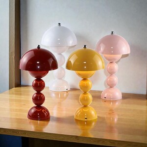 May include: Four modern table lamps in red, white, yellow, and pink. Each lamp has a mushroom-shaped shade and a spherical base. The lamps are displayed on a wooden surface. Each lamp has a silver-colored top.