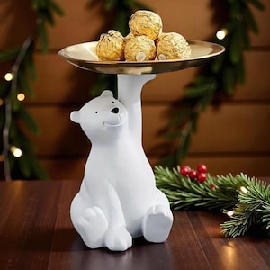 Pode incluir: Uma estatueta de urso polar branco segurando uma bandeja dourada com vários chocolates embrulhados em ouro. O urso está sentado com as pernas estendidas, e a bandeja é segurada acima de sua cabeça. O fundo inclui decorações de Natal.