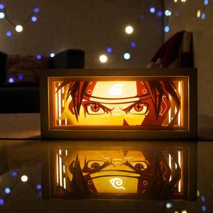Pu&ograve; includere: Un'opera d'arte in carta illuminata, di forma rettangolare, raffigurante il volto di un personaggio anime con capelli e occhi rossi. L'opera &egrave; racchiusa in una cornice di legno chiaro, con un riflesso sottostante. Lo sfondo &egrave; scuro, con luci a catena.