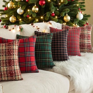 Op de afbeelding: Een verzameling geruite kussens in rood, groen en zwart, gerangschikt op een witte bank met een imitatiebont plaid. Een kerstboom met ornamenten en lichtjes staat op de achtergrond en creëert een feestelijke vakantiescène.