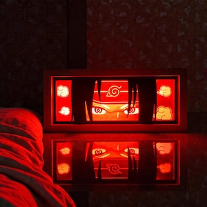 Pu&ograve; includere: Una luce notturna rettangolare rossa a tema anime, con gli occhi e la fascia di un personaggio. Il design include nuvole stilizzate e un simbolo, con un riflesso a specchio. La cornice &egrave; marrone caldo.