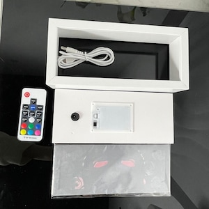 Pu&ograve; includere: Una scatola luminosa a LED bianca con interno nero, un cavo USB e un telecomando. Il telecomando ha pi&ugrave; opzioni di colore. La scatola luminosa ha un vano batteria e un piccolo pulsante nero. Un'immagine avvolta in plastica &egrave; in basso.