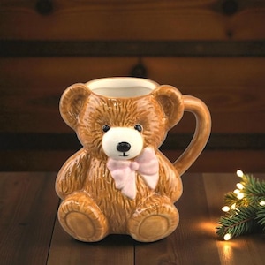 May include: A brown ceramic teddy bear mug with a pink bow. The mug has a handle and is designed to look like a teddy bear, with a white face, black eyes, and a black nose. The mug is sitting on a wooden surface.